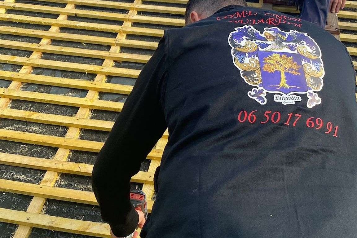 Un couvreur Dujardin, le blason de l'entreprise dans le dos, au travail sur le lattage d'une toiture